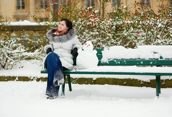 Ragazza seduta sulla panchina con divertente pupazzo di neve — Foto Stock