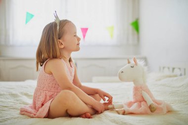 Pembe elbiseli ve prenses gibi giyinmiş tek boynuzlu atlarla oynayan sevimli küçük bir kız. Çocuk yumuşak oyuncakla eğleniyor. Çocuk doldurulmuş oyuncakla oynuyor