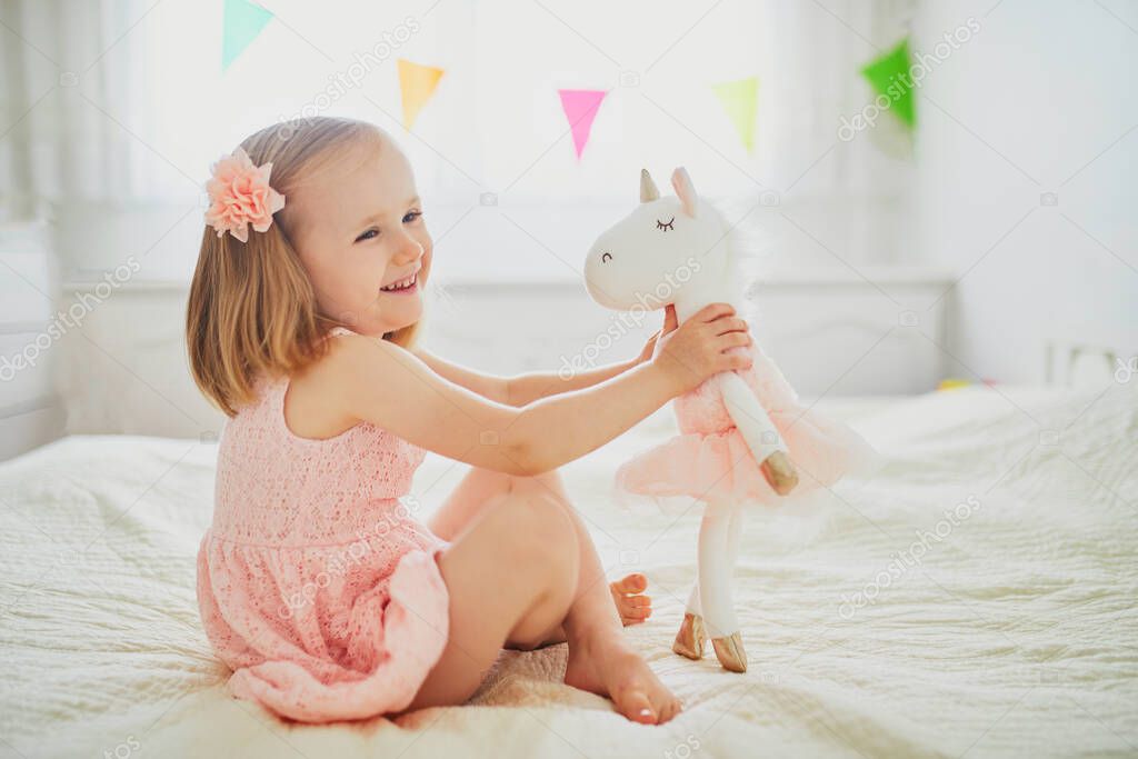 Adorable little girl in pink dress playing with unicorn. Kid having fun with soft toy. Child playing with stuffed toy