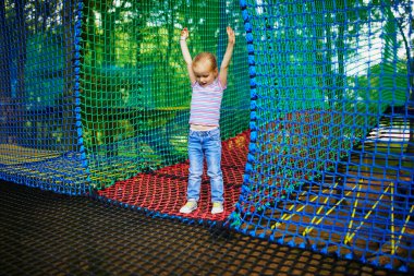 Küçük sevimli kız macera parkında eğleniyor. Ağacın tepesindeki trambolinde bir çocuk. Küçük çocuklar için açık hava aktiviteleri