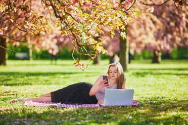Güzel genç kız kiraz çiçeği mevsiminde parkta dizüstü bilgisayarıyla çalışıyor. Paris, Fransa yakınlarındaki ünlü Sceaux Parkı 'nda genç bir kadın. Serbest çalışma, uzaklık öğrenme veya uzaktan çalışma kavramı