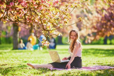 Güzel genç kız kiraz çiçeği mevsiminde parkta dizüstü bilgisayarıyla çalışıyor. Paris, Fransa yakınlarındaki ünlü Sceaux Parkı 'nda genç bir kadın. Serbest çalışma, uzaklık öğrenme veya uzaktan çalışma kavramı