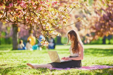 Güzel genç kız kiraz çiçeği mevsiminde parkta dizüstü bilgisayarıyla çalışıyor. Paris, Fransa yakınlarındaki ünlü Sceaux Parkı 'nda genç bir kadın. Serbest çalışma, uzaklık öğrenme veya uzaktan çalışma kavramı