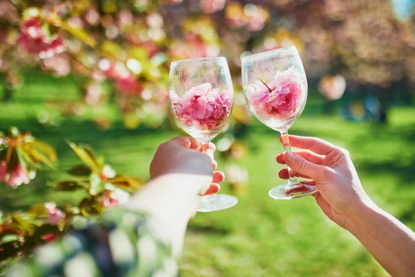春の日にはピンクの桜の花びらでいっぱいのワイングラスを持っている2人の女性の手 フランス パリ近郊の有名なスコー公園の桜の季節 — ストック写真