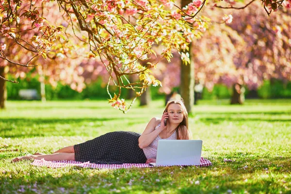 樱花盛开的季节 漂亮的小女孩在公园的笔记本电脑上工作 在法国巴黎附近著名的Sceaux公园里的年轻女子 自由职业 远程学习或远程工作概念 — 图库照片