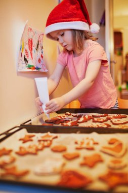 Kırmızı Noel Baba şapkalı sevimli anaokulu kızı Noel kurabiyesi pişiriyor. Evde çocuklarla mevsimlik tatilleri kutluyoruz.