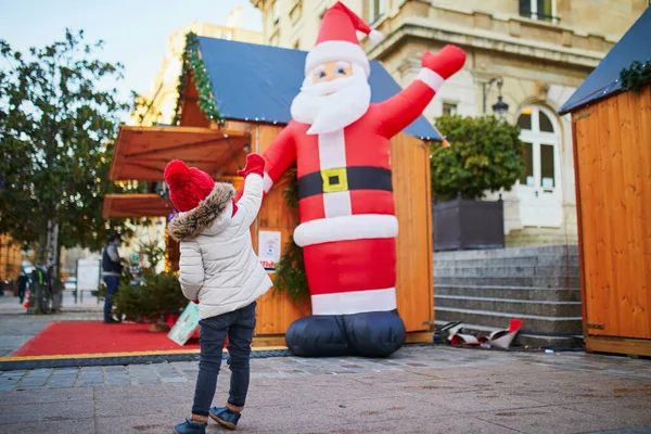 幸せな陽気な就学前の女の子で赤い帽子挨拶人工サンタクロース上のクリスマスマーケットでパリ フランス — ストック写真