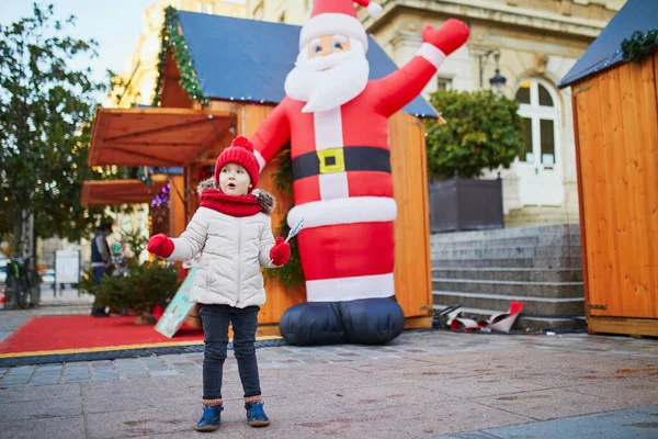 幸せな陽気な就学前の女の子で赤い帽子挨拶人工サンタクロース上のクリスマスマーケットでパリ フランス — ストック写真