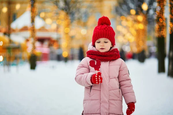 幸せな陽気な就学前の女の子で赤い雪と帽子クリスマスマーケットで楽しみ 鶏形の砂糖ロリポップを食べる — ストック写真