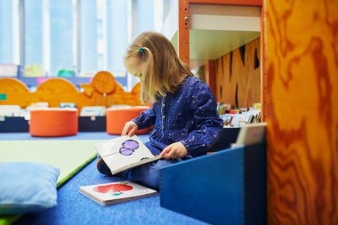 4 yaşında bir kız kütüphanede yerde oturuyor ve kitap okuyor. Okuma ve öğrenme konsepti