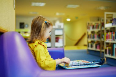 4 yaşında bir kız kütüphanede yerde oturuyor ve kitap okuyor. Okuma ve öğrenme konsepti