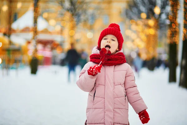 幸せな陽気な就学前の女の子で赤い雪と帽子クリスマスマーケットで楽しみ 鶏形の砂糖ロリポップを食べる — ストック写真