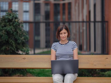 Dizüstü bilgisayarda çalışan öğrenci şehir caddesinde bankta oturuyor..