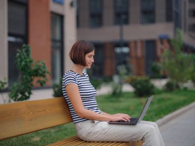 Dizüstü bilgisayarda çalışan öğrenci şehir caddesinde bankta oturuyor..