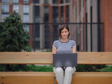 Dizüstü bilgisayarda çalışan öğrenci şehir caddesinde bankta oturuyor..