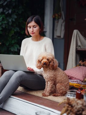 Ev terasında çalışan Asyalı bir kadın, sonbaharın tadını çıkaran köpekle rahat bir şekilde oturuyor, sosyal ağda dizüstü bilgisayarla mesaj yazan bir kadın etkisinde. çalışma ve öğrenim konsepti