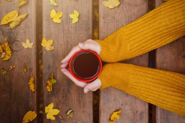 Bir fincan sıcak çay, bayanların elinde, sonbahar arkaplanındaki ahşap masada yapraklarla duruyor. Sıcak içecek konsepti.