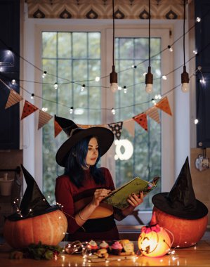 Tüm kutsal arifeler. Cadı kostümlü ve şapka takan kadın kitap okuyor. Masaya balkabağı oyuluyor. Karanlık odada Cadılar Bayramı süslemeleri yapılıyor.