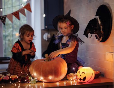 Cadılar bayramı kutlaması sırasında parlayan balkabağı feneriyle oynayan cadı kostümlü küçük kız kardeşler karanlık mutfak odasında, masada heyecanlı çocuklar ve balkabakları