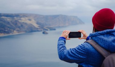 Bir kadın akıllı telefondan denizin fotoğrafını çeker, dişi soğuk mevsimde kıyı boyunca seyahat eder. Açık havada. Bakış açısı. Kapat.