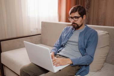 Evde dizüstü bilgisayar kullanan yakışıklı bir genç. Öğrenci erkekler odasında. Çevrimiçi alışveriş, ev işi, serbest çalışma, çevrimiçi öğrenme, öğrenim kavramı. Uzaklık eğitimi