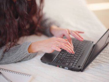 Yatakta dizüstü bilgisayar kullanan, rahat gri örülü kazaklı bir kadın.