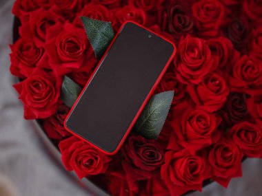 Akıllı telefon, kırmızı gül buketi ve hafif yatakta taç yaprakları, üst manzara. Anneler günü, sevgililer günü, doğum günü, seçici odak