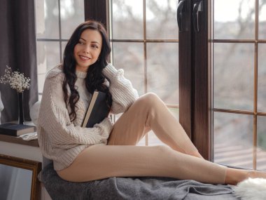 Evdeki pencerenin yanında kitap okuyan güzel genç bir kadın. Rahatlamak için boş zaman. Pencerenin yanında mola veren mutlu bayan, boşluğu kopyala