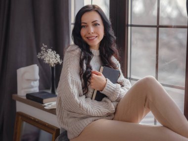 Evdeki pencerenin yanında kitap okuyan güzel genç bir kadın. Rahatlamak için boş zaman. Pencerenin yanında mola veren mutlu bayan, boşluğu kopyala