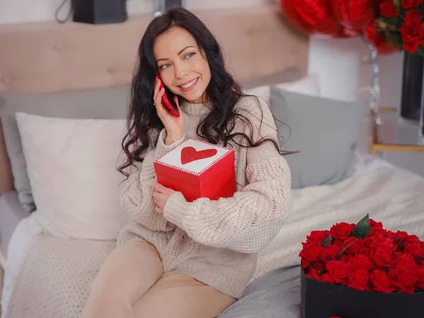 Gülümseyen mutlu kadın, hediye kutusuyla telefonda sevdikleriyle konuşuyor, yatakta oturup kameraya bakıyor. Sevgililer Günü 'nün kutlanması, düğün ve aşk günü, kadınlar günü. Sabah aşk yatak odası.