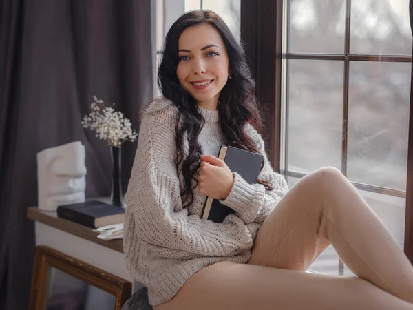 Evdeki pencerenin yanında kitap okuyan güzel genç bir kadın. Rahatlamak için boş zaman. Pencerenin yanında mola veren mutlu bayan, boşluğu kopyala