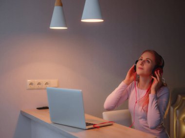 Oturma odasında dizüstü bilgisayarla çalışan çekici genç bir kadın. Genç bayanın favori şarkısı kulaklıkla dinleniyor ve ev bilgisayarında çalışırken şarkı söylüyor..