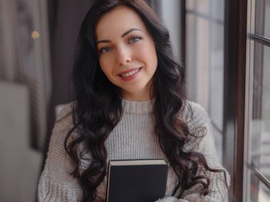 Evdeki pencerenin yanında kitap okuyan güzel genç bir kadın. Rahatlamak için boş zaman. Pencerenin yanında mola veren mutlu bayan, boşluğu kopyala