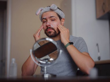 Men's beauty idea. Young handsome man in the kitchen in hygiene concept . headband, mirror on the table, cleanser for skin imperfections