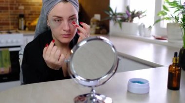 Gözler için yamalar. Mutfakta, kafasında havluyla çekici bir kadın gözlerinin altındaki deriye şeffaf yamalar yapıştırıyor. Hijyen, güzellik uzmanı, makyaj, yüz bakımı konsepti.