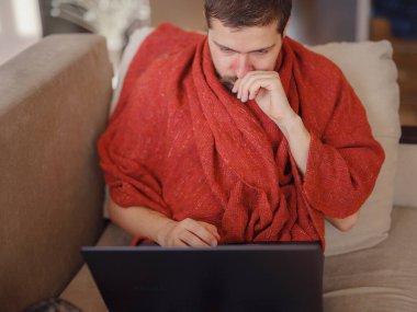Sıcak battaniyeye sarılı bir adam dizüstü bilgisayara bakıyor, evdeki kanepede oturuyor, boş bir alan. Sinüzit hastalığı, soğuk algınlığı ve grip belirtileri. Evde uzaktan çalışıyor..