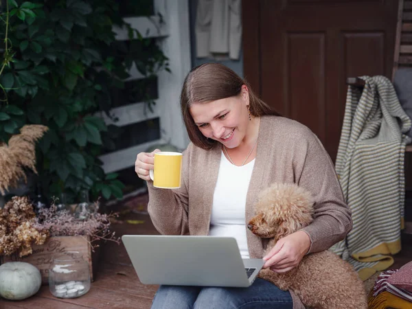 middle aged caucasian woman in brown cardigan sits on terrace of house with her beige poodle with laptop working outdoors in garden, home office , Education, modern lifestyle and leisure concept.