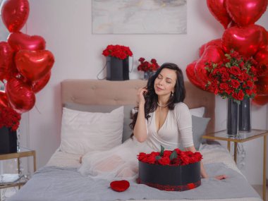 Beyaz elbiseli genç bir kadın yatakta oturuyor. Sabah aşkı yatak odası. Yıldönümü, mutlu yıllar, düğün, aşk günü, kadınlar günü, sevgililer günü ve olumlu duygular.