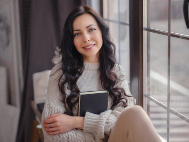 Evdeki pencerenin yanında kitap okuyan güzel genç bir kadın. Rahatlamak için boş zaman. Pencerenin yanında mola veren mutlu bayan, boşluğu kopyala