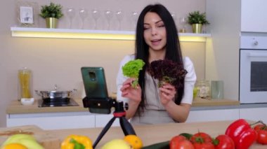 Professional beautiful happy young woman is blogging for her kitchen channel about healthy living in kitchen of her home and looking on smartphone on tripod