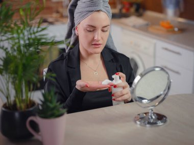 Enjoying flawless skin . Peaceful lady being satisfied with condition of her skin during procedures in kitchen. Beautiful caucasian woman is cleaning face with Cleansing foam looking in mirror.