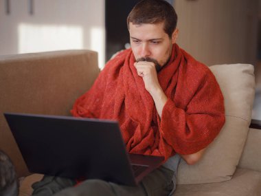 Sıcak battaniyeye sarılı bir adam dizüstü bilgisayara bakıyor, evdeki kanepede oturuyor, boş bir alan. Sinüzit hastalığı, soğuk algınlığı ve grip belirtileri. Evde uzaktan çalışıyor..