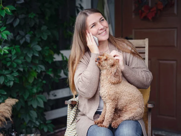 middle aged caucasian woman in brown cardigan sits on terrace of house with her beige poodle and emotionally speaks with friend via smartphone. morning good and bad news, autumn lifestyle concept