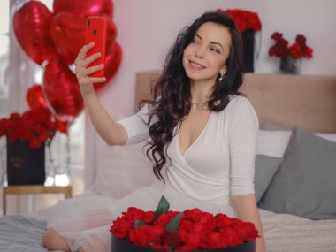Beyaz elbiseli genç bir kadın akıllı telefonuyla selfie çekiyor ve yatakta oturuyor. Sabah aşkı yatak odası. Yıldönümü, mutlu yıllar, düğün, aşk günü, kadınlar günü, sevgililer günü ve olumlu duygular.
