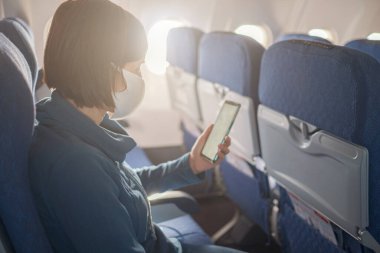 Genç ve güzel bir kadın uçuş sırasında uçağın penceresinde oturuyor. Covid-19 salgınından sonra yeni normal seyahat. Akıllı telefon monitörü ekranını, maketini göster