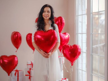 Beyaz elbiseli güzel, mutlu genç bir kadın. Tatil partisi. Kırmızı kalp şeklinde balonlarla poz veren neşeli manken, eğlenen, Sevgililer Günü 'nü kutlayan. Aşkın sembolü