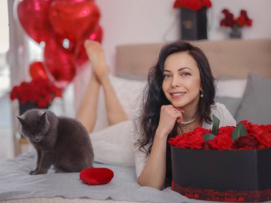 Beyaz elbiseli genç bir kadın gri kediyle yatakta oturuyor. Sabah aşkı yatak odası. Yıldönümü, mutlu yıllar, düğün, aşk günü, kadınlar günü, sevgililer günü ve olumlu duygular.
