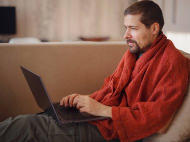 Sıcak battaniyeye sarılı bir adam dizüstü bilgisayara bakıyor, evdeki kanepede oturuyor, boş bir alan. Sinüzit hastalığı, soğuk algınlığı ve grip belirtileri. Evde uzaktan çalışıyor..