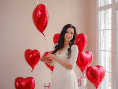 Beyaz elbiseli güzel, mutlu genç bir kadın. Tatil partisi. Kırmızı kalp şeklinde balonlarla poz veren neşeli manken, eğlenen, Sevgililer Günü 'nü kutlayan. Aşkın sembolü