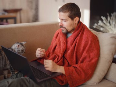 Sıcak battaniyeye sarılı bir adam dizüstü bilgisayara bakıyor, evdeki kanepede oturuyor, boş bir alan. Sinüzit hastalığı, soğuk algınlığı ve grip belirtileri. Evde uzaktan çalışıyor..